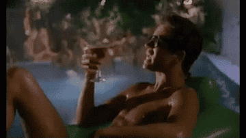 George Michael sipping a drink while lounging in a pool with his shirt off, then pouring the drink out