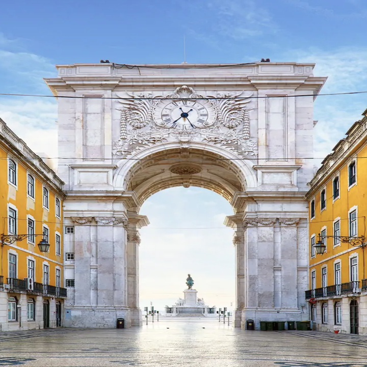A shot of a large archway in Lisbon with a clockface and surrounded by bright, yellow buildings and no tourists in front of it