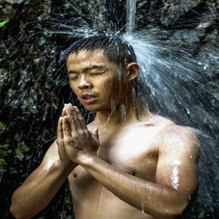 A shirtless man holds his hands in prayer position as a stream of water from a waterfalls rains on his head