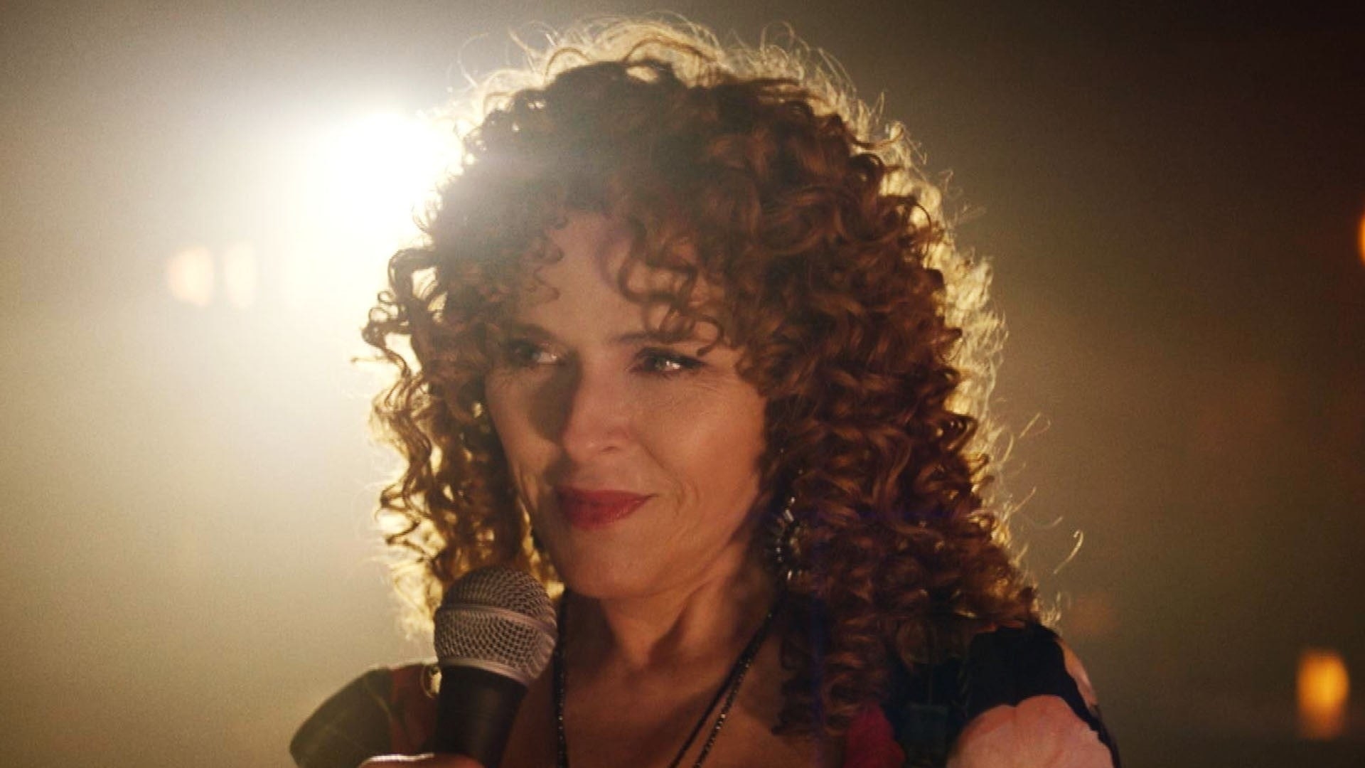 Bernadette Peters smiling warmly about to sing into a mic on a dim stage