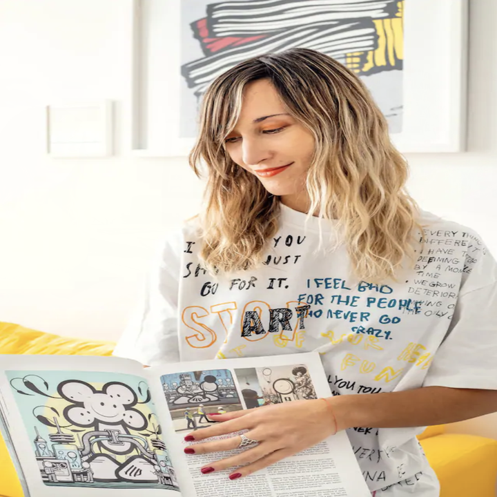 A woman in a graffitied t shirt smiles and holds an art book to the camera