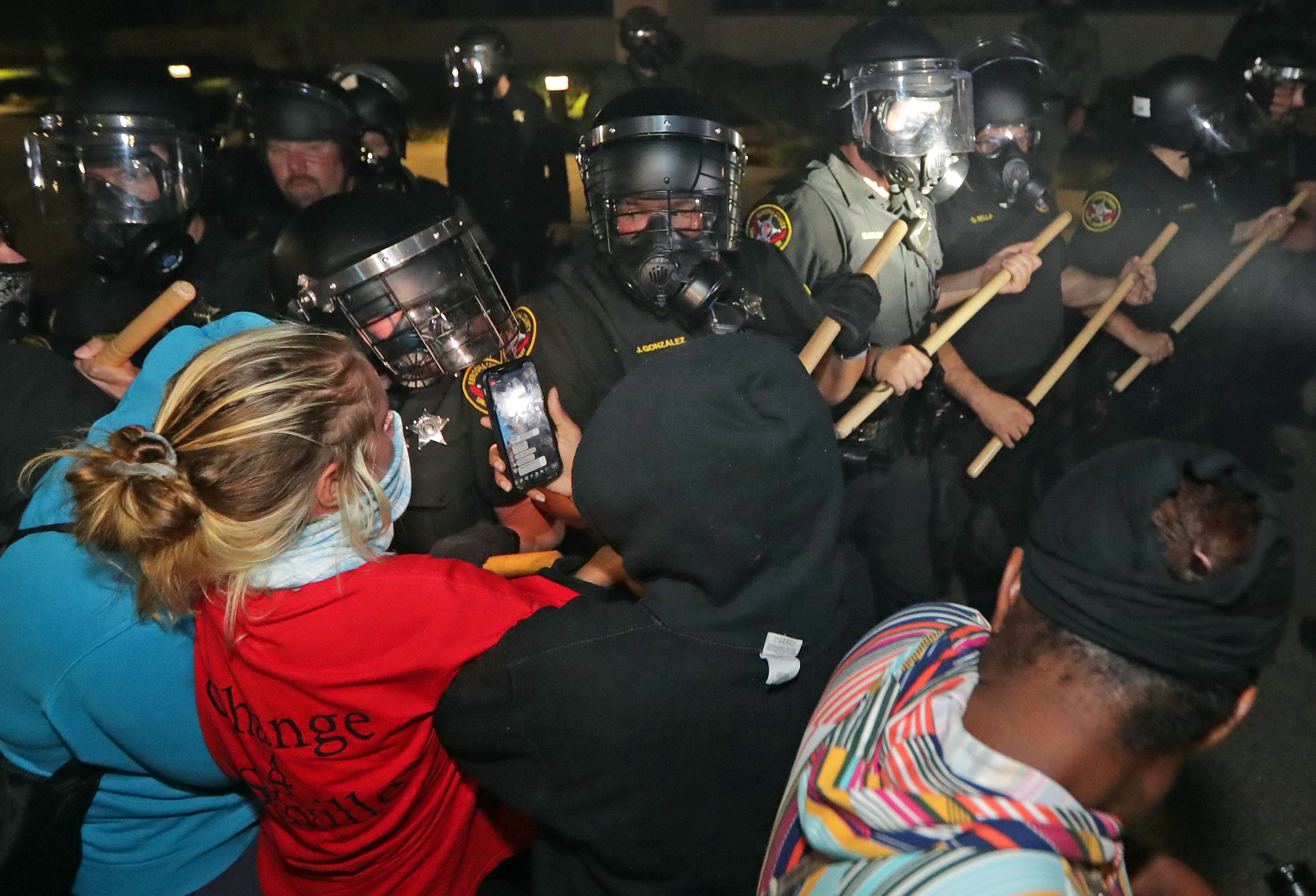 Officers with face shields and batons confront protesters, one of whom is filming on their smartphone 