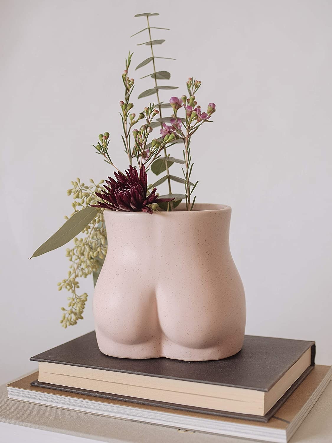 A vase that&#x27;s shaped like a butt in light pink with a wide open top with flowers in it