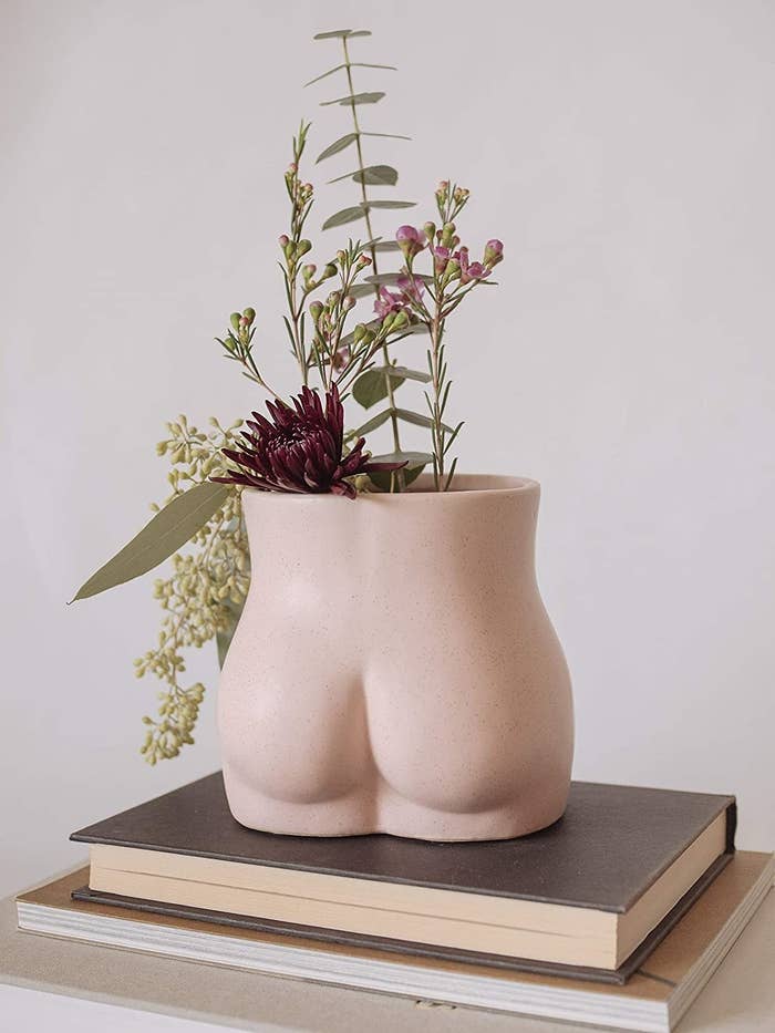 A vase that's shaped like a butt in light pink with a wide open top with flowers in it