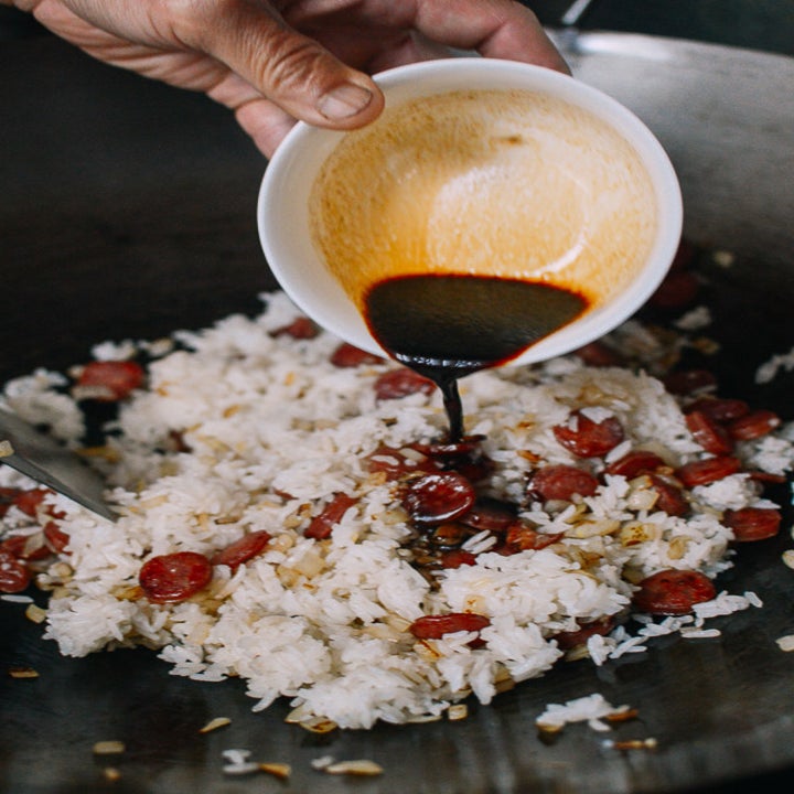 Someone pouring soy sauce from a small bowl onto a skillet of sausage and white rice.