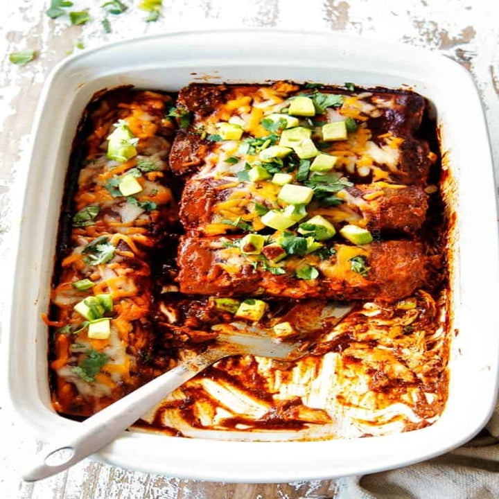 A baking pan filled with chicken enchiladas topped with enchilada sauce, cheese, and diced avocado.
