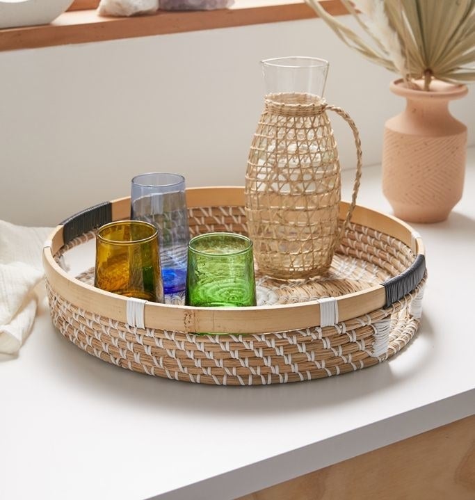 Woven seagrass serving tray with cutout side handles and assorted glasses in it