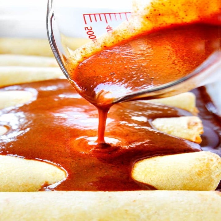 Red enchilada sauce being poured onto tortillas rolled up with chicken.