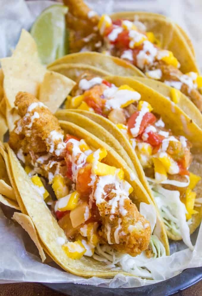 Three crispy fish tacos in tortillas with cabbage, mango and tomato salsa, and crema on top.