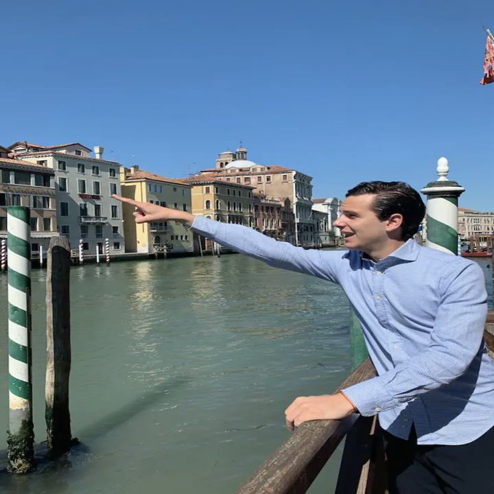 A man stands on a dock over a canal and points excitedly towards a row of buildings across the water