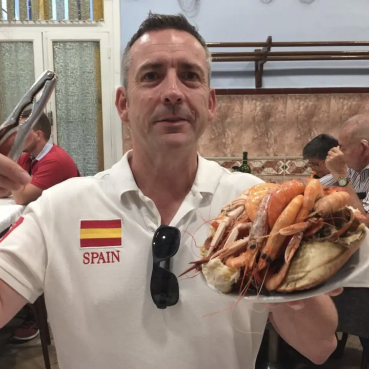 A man in a shirt decorated with a Spanish flag sits in a restaurant and holds up a plate full of seafood in one hand and tongs in the other