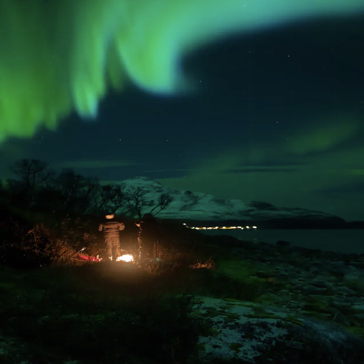 Bright neon lights swirl in the night sky above the figure of a man in front of a fire and snowy mountains in the background
