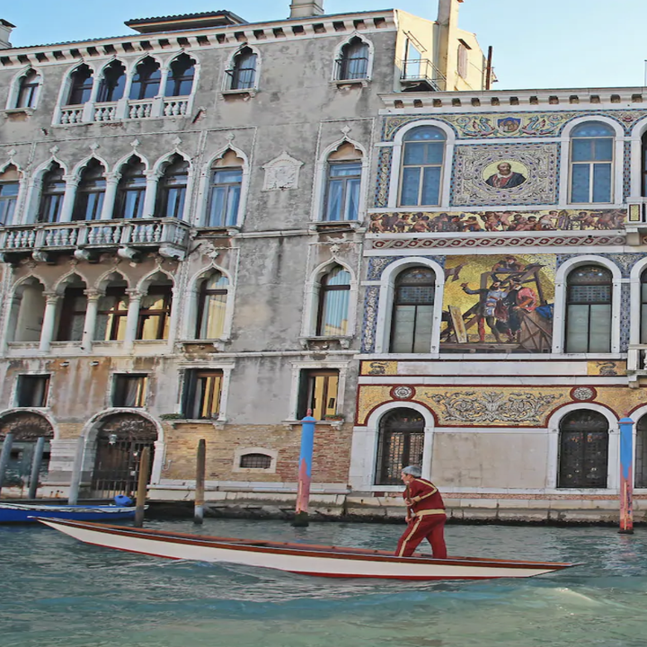 A man rows an empty gondola across a canal in front of intricately decorated buildings