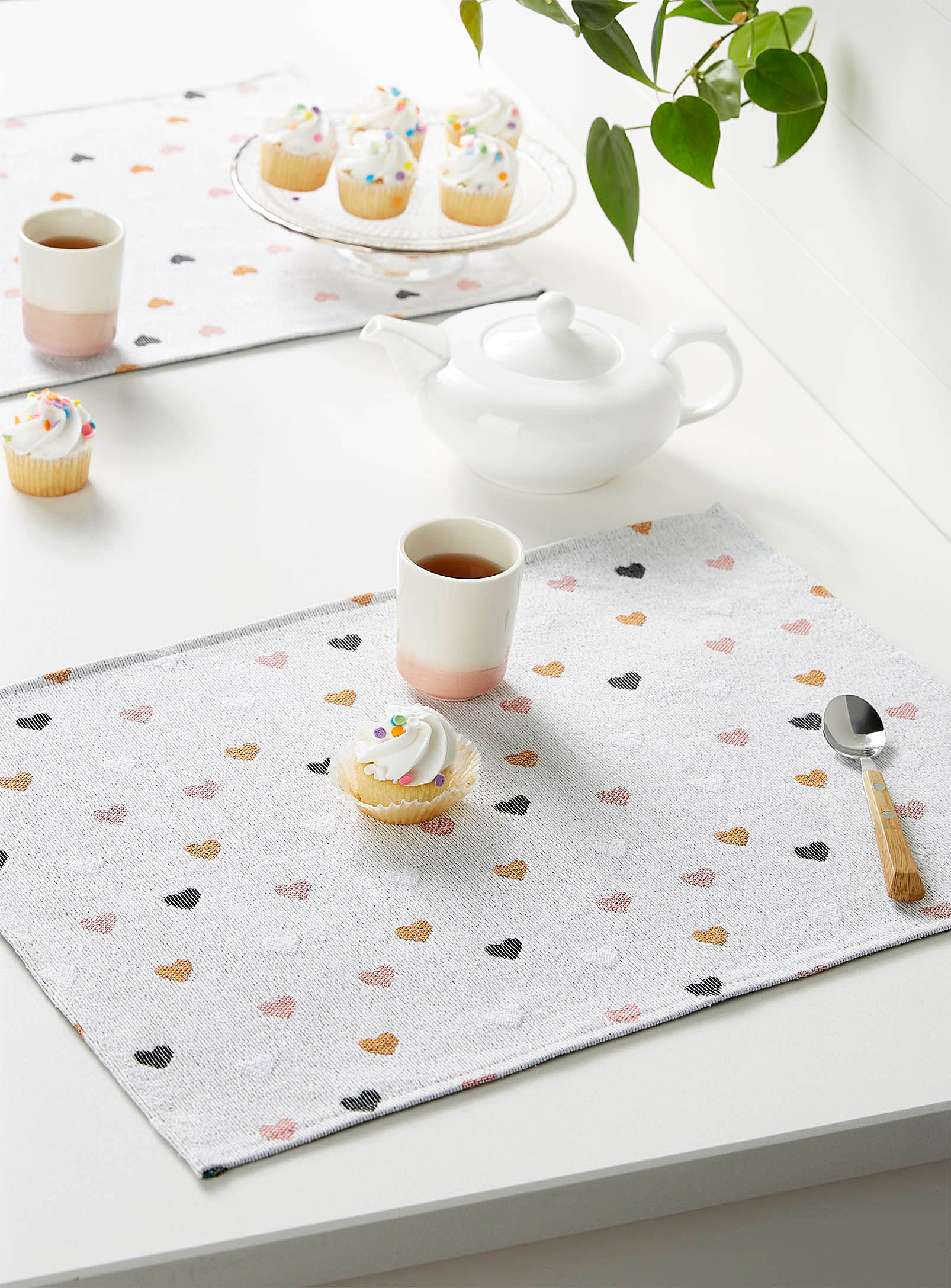 A rectangular cloth placemat on a table with a cupcake, a cup and a spoon on it