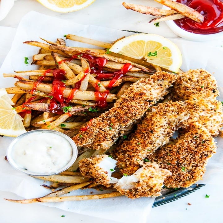 A plate with baked fish sticks, tartar sauce on the side and French fries covered in Ketchup.