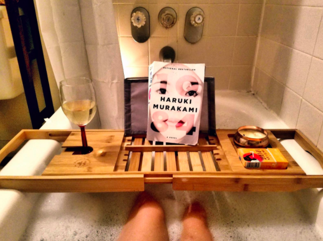 Reviewer's light wood bath caddy with a glass of white wine, a book, and candle over their bath tub