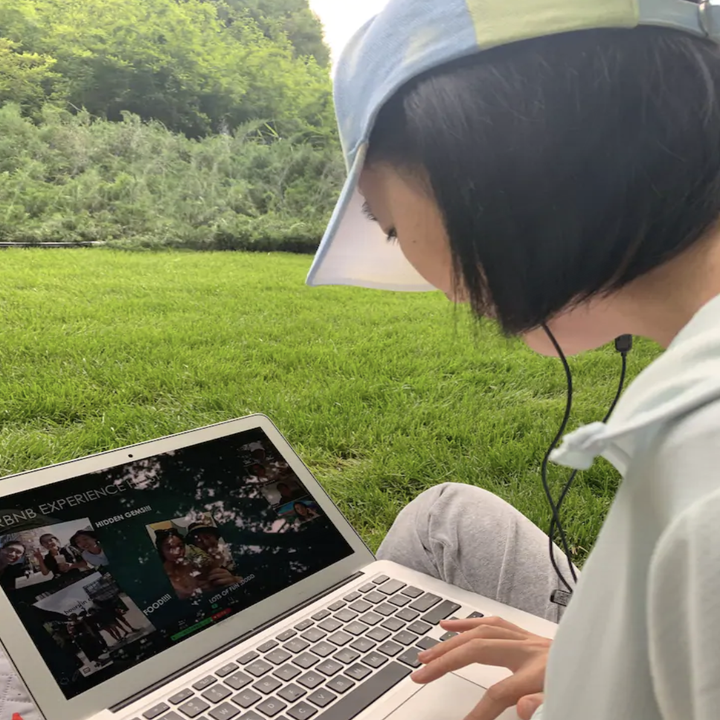 A woman's side profile featuring a short bob and baseball hat as she sits on grass and looks down at her laptop