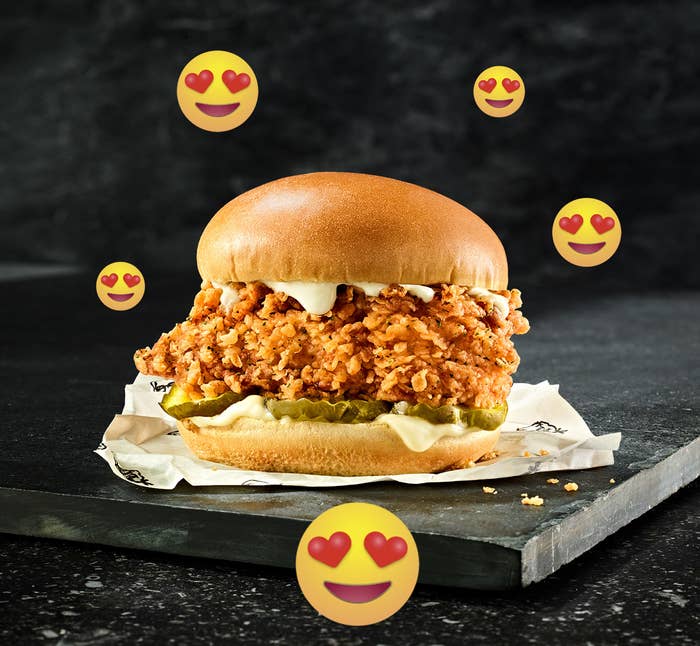The Famous Chicken Chicken Sandwich is sitting on a piece of slate against a dark background. Emojis with hearts in their eyes are scattered across the image.