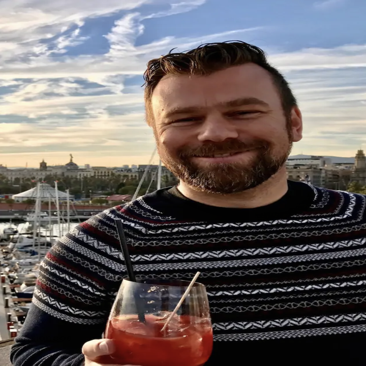 A man smiles at the camera as the sun sets behind him and he holds a cocktail out to the camera