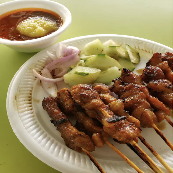 A paper plate holds skewers of meat, fresh cucumber, and onion with a bowl of spicy-looking sauce on the side