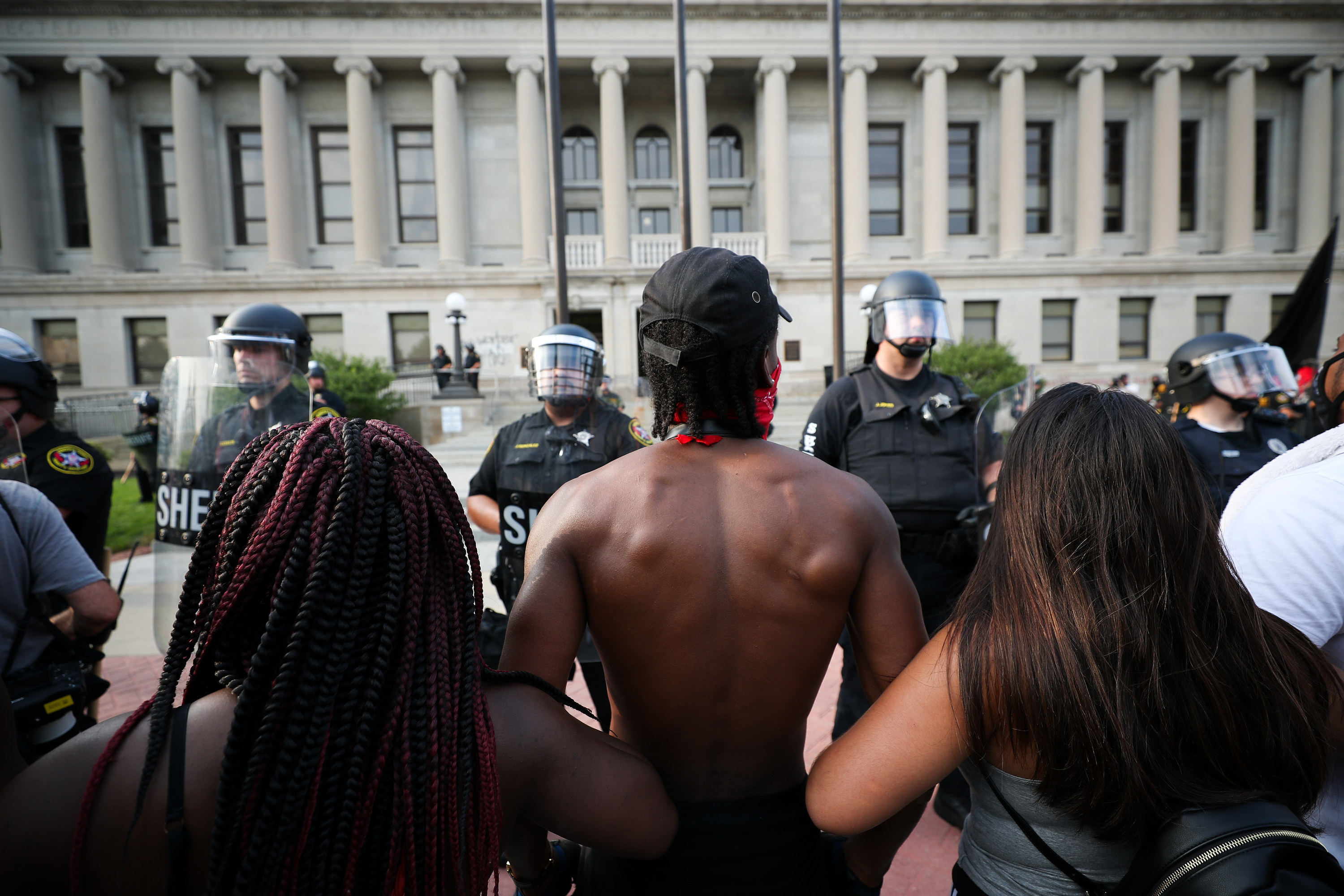 A black man from behind with his arms linked to other protesters faces off with police in riot gear in front of the courthouse