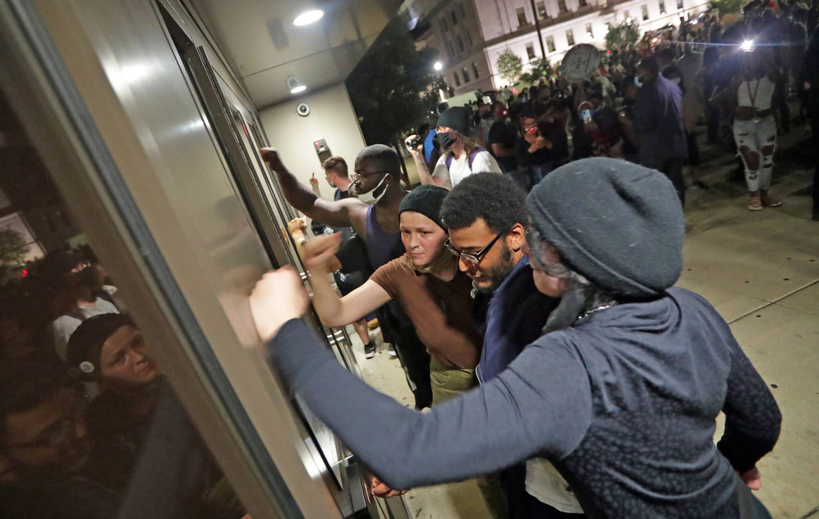 Three people pound on a door with a larger crowd behind them 