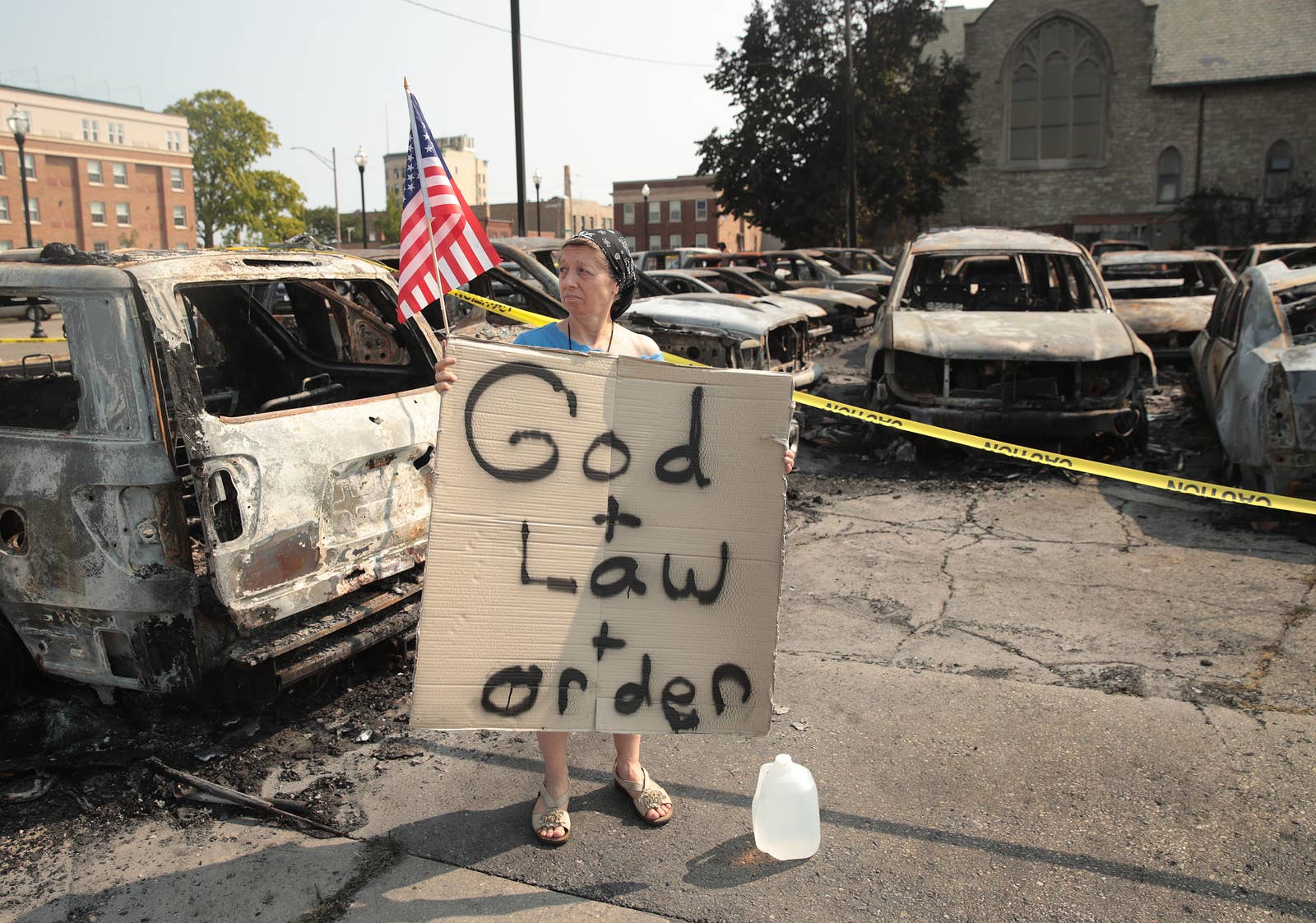 A woman holds a small American flag and a cardboard sign that says god + law + order