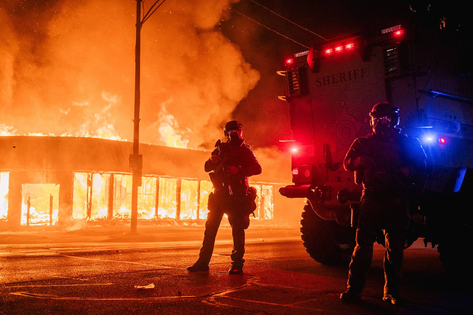 Two police in riot gear stand in front of a burning building 