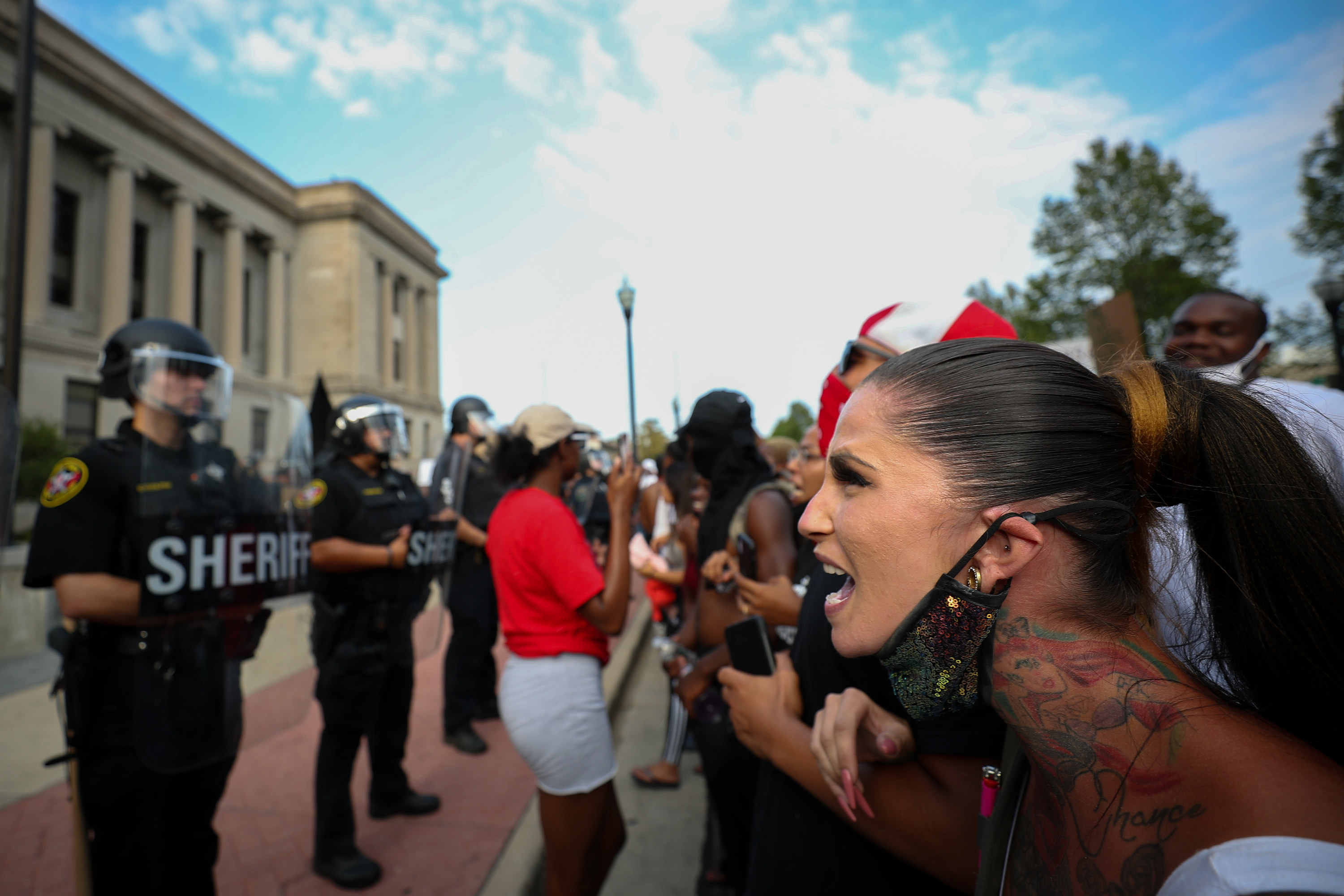A white woman yells while linking arms with other protesters in front of a line of police in riot gear
