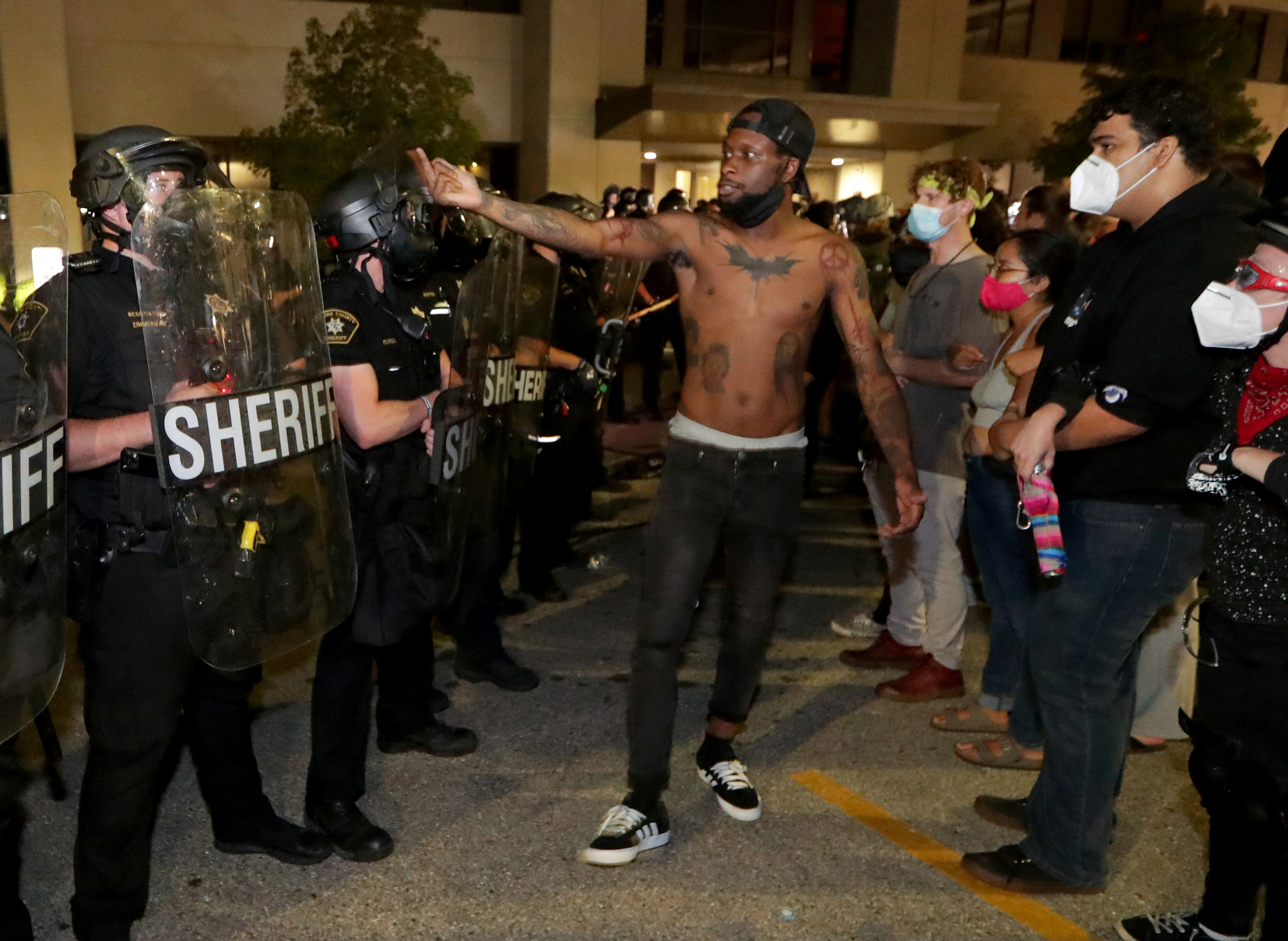 A shirtless Black man uses gestures with his pointer and middle finger raised to a line of police with a crowd next to him 