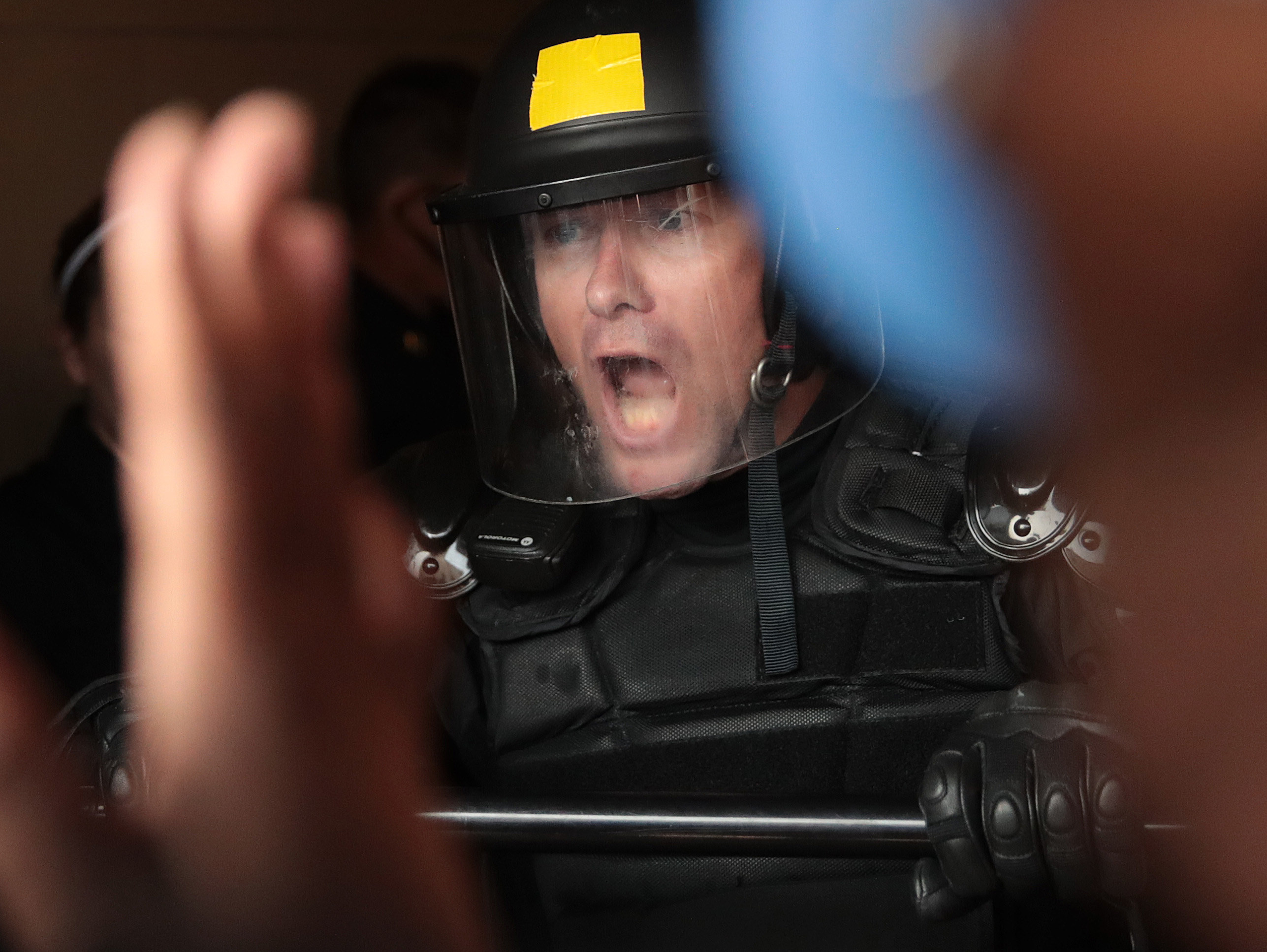 A white police officer in riot gear and a face shield yells