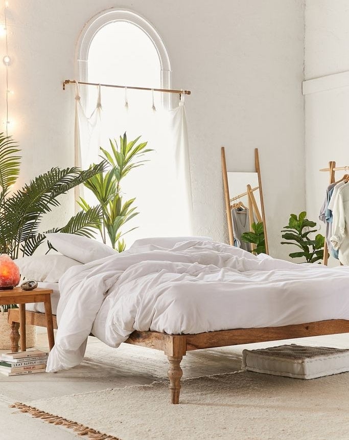 A wooden bed platform with no headboard holding a bed with only a white comforter and white pillows. The bed sits near an arched window, while the rest of the room has a small wooden table, a rug, a wooden mirror, and plant situated around the bed.