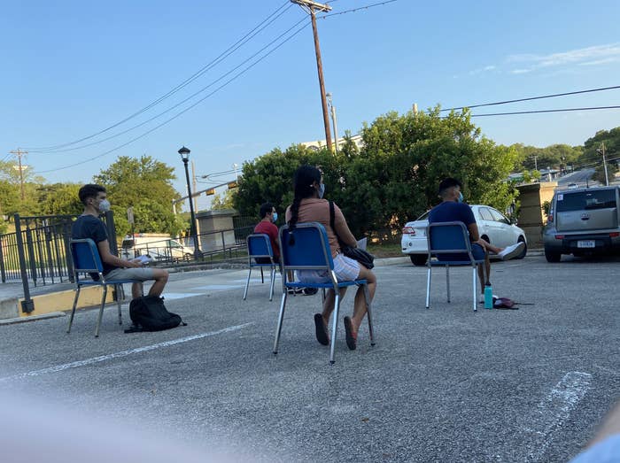 Four students in face masks sit in plastic chairs spaced 6 feet apart on an asphalt parking lot.