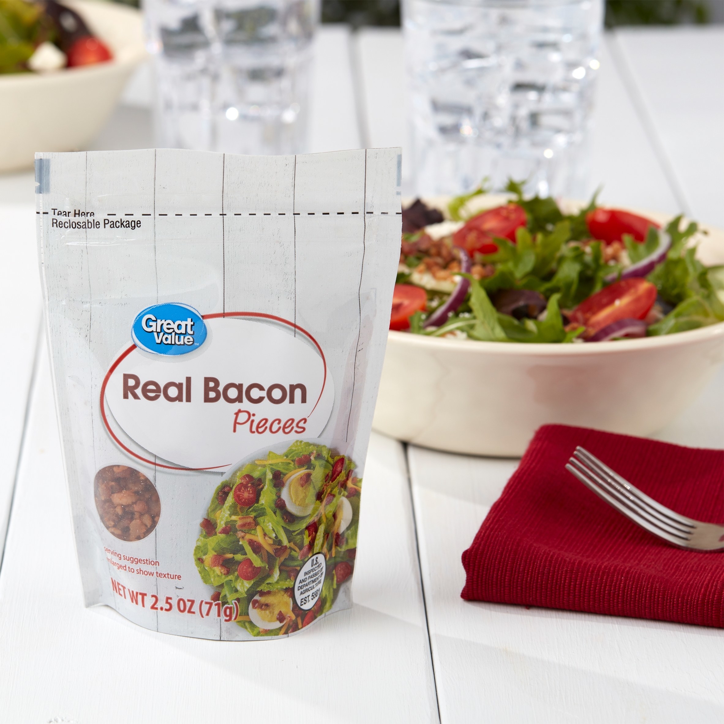 Pack of bacon pieces on a table next to a salad, napkin, and fork
