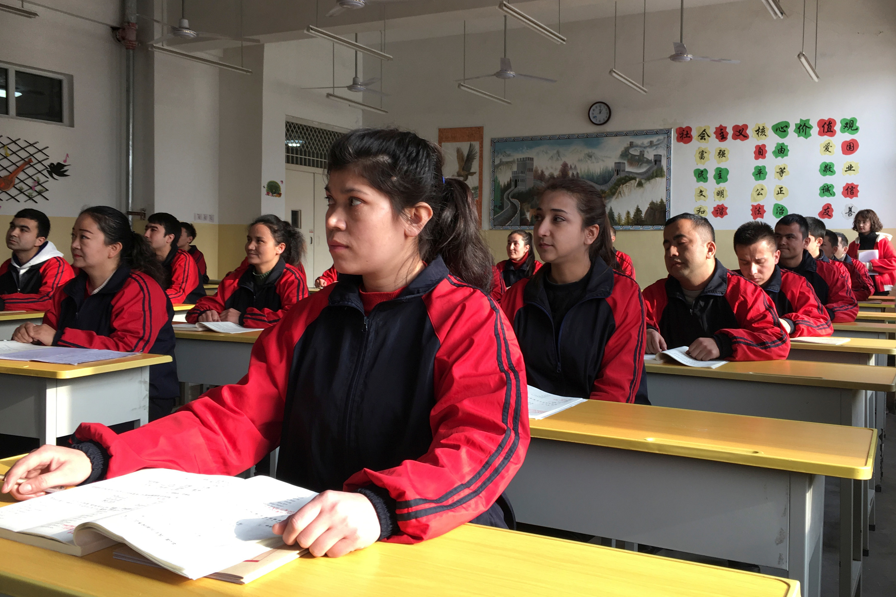 Adult students in matching jumpsuits sit in rows of desks