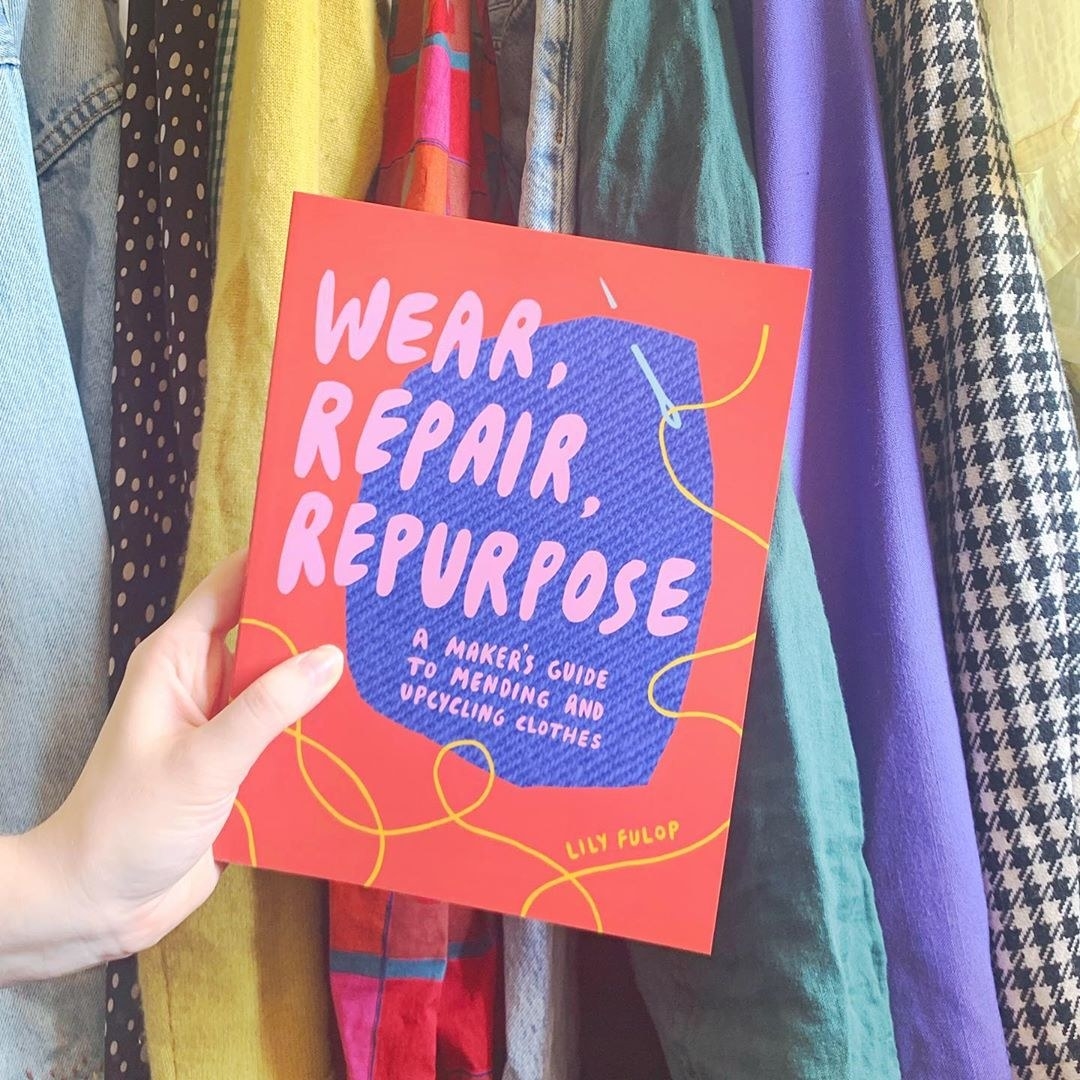 A person holding a book called Wear, Repair, Repurpose in front of a rack of clothing