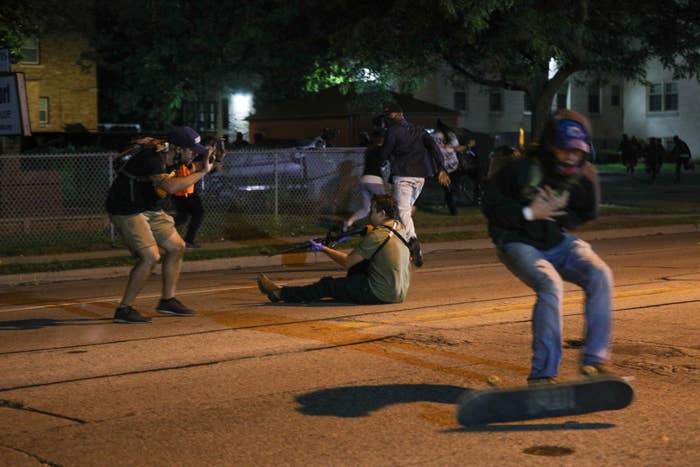 Someone sits on the street holding a long gun as protesters react and scatter, one in the foreground falls from a skateboard