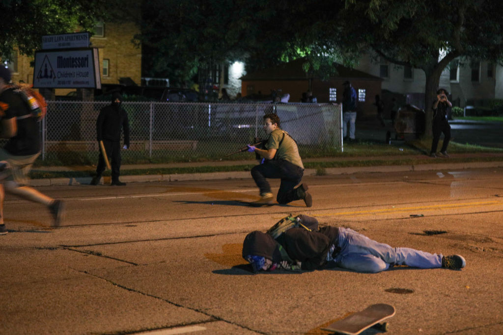 A person lies on the ground near their skateboard, behind them, a man kneels with a long gun pointed in a different direction