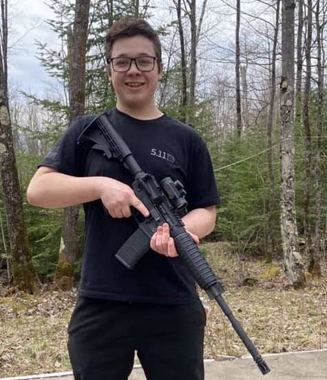 A teenager dressed in a black T-shirt and pants smiles and holds a large gun