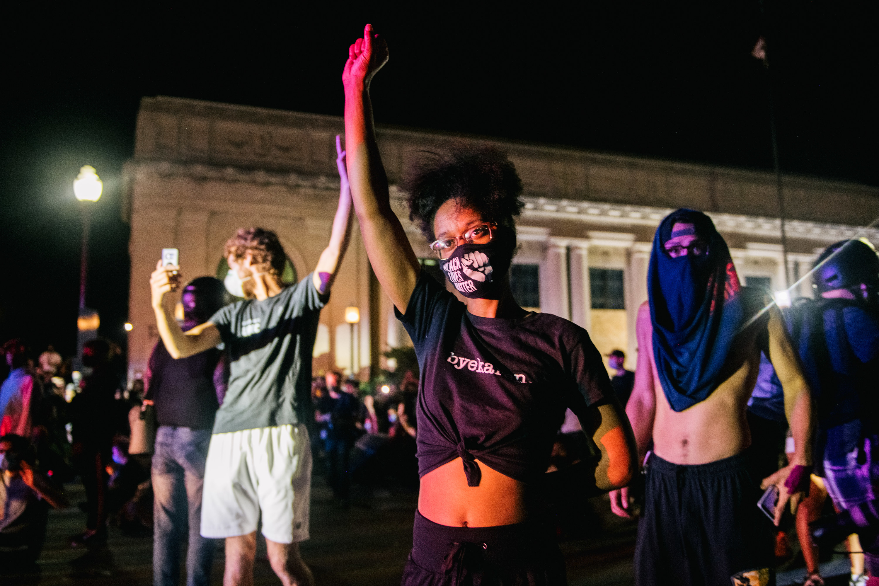 A Black woman, wearing a tied-up black T-shirt and face mask that reads &quot;Black Lives Matter,&quot; holds up her fist and looks into the camera