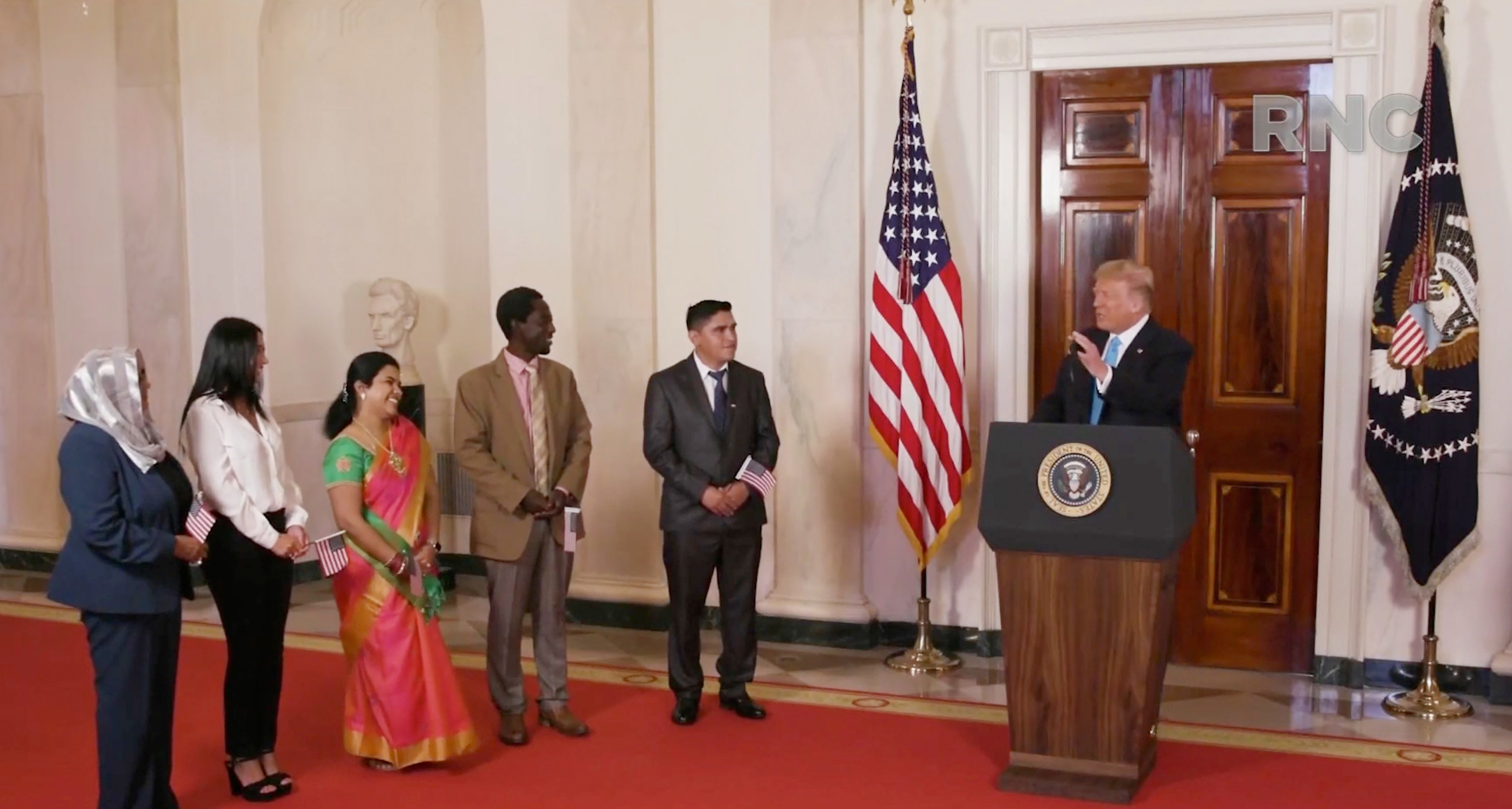 Donald Trump stands behind a lectern and speaks toward five other people standing nearby