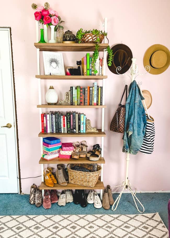 Reviewer pic of the six-tier bookshelf with light brown wood shelves and white rods attached to the wall with books and trinks on the top four shelves and assorted shoes and accessories on the bottom shelves