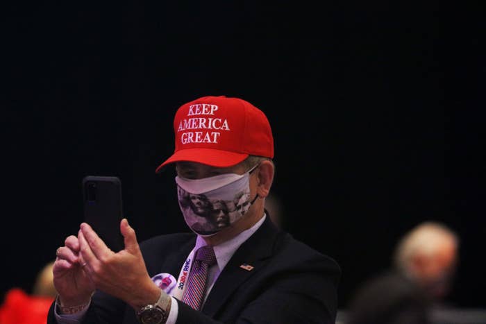 A person wearing a Mount Rushmore face mask and a MAGA hat holds his smartphone up