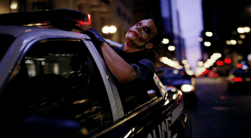 The Joker sticks his head out of a police car window as it zooms through the city.