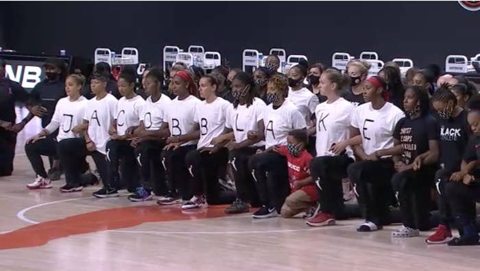 WNBA players kneel in a row, each wearing a white T-shirt with a letter on it, spelling out "Jacob Blake"