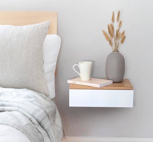 Rectangle white wood drawer with a light brown wood top attached to the wall next to the bed with a book, coffee cup, and vase on it