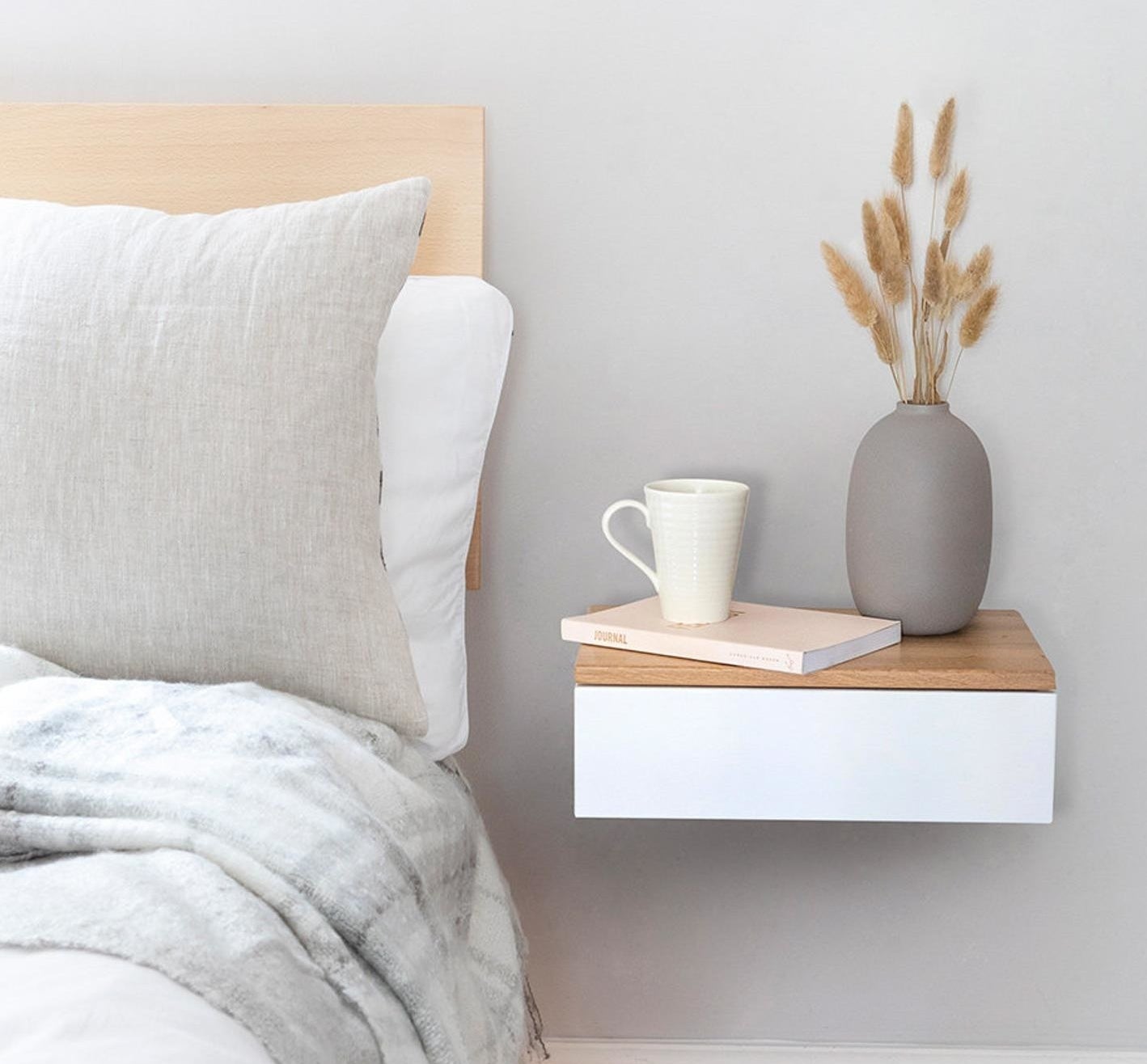 Rectangle white wood drawer with a light brown wood top attached to the wall next to the bed with a book, coffee cup, and vase on it