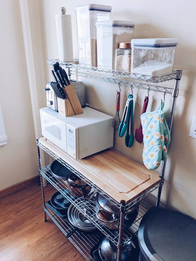 Reviewer photo of the metal baker's rack with two shelves on bottom and high shelf on top filled with kitchen products and a microwave