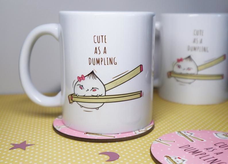 A mug with a dumpling with a cute face being squeezed by chopsticks that says cute as a dumpling