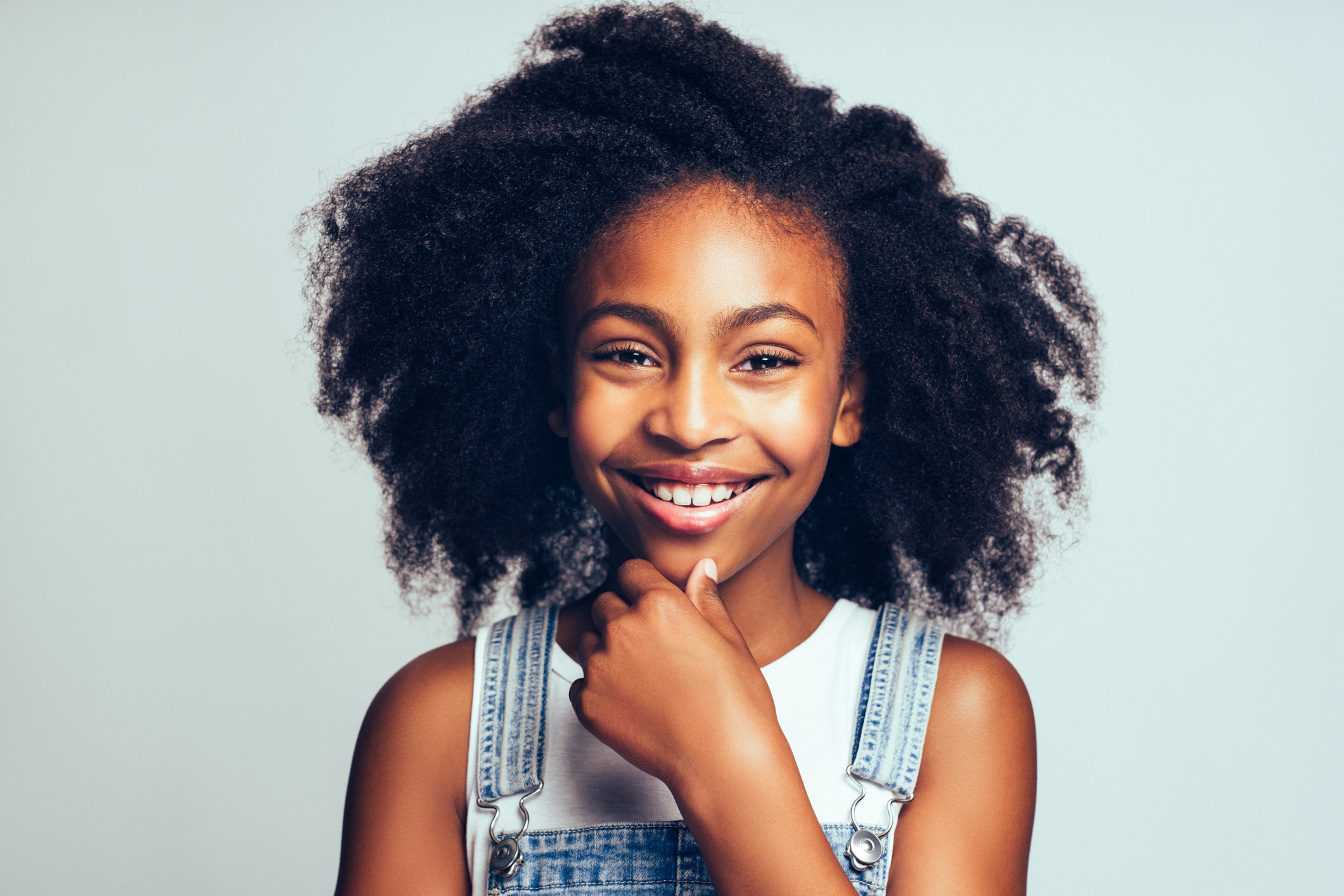 Girl smiling and wearing overalls standing with her hand on her chin against a gray background